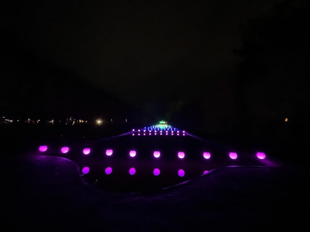 A series of glowing orbs arranged in a symmetrical pattern creating a pathway that appears to extend into the distance. The orbs are primarily purple, with a few shifting into pink and blue tones as they recede. The scene is set against a dark background, emphasizing the luminous quality of the lights.