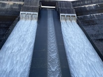 A large concrete structure with three spillways releasing a significant amount of water. The two outer spillways have heavier flows, while the middle spillway has a lighter, more concentrated stream. The concrete is dark and weathered, with white, frothy water cascading down.