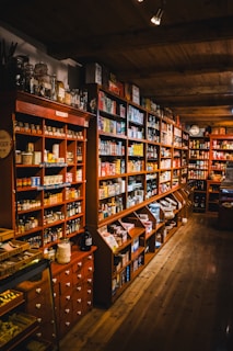 An interior of a vintage-style store is filled with shelves stocked with an assortment of goods, including packaged foods and glass jars. The room features warm wooden shelving and flooring, creating a cozy and nostalgic atmosphere. Soft lighting enhances the rustic charm of the setup.