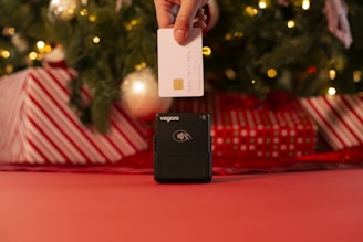 A hand is holding a white credit card above a contactless card reader. The background features a decorated Christmas tree with lights and wrapped gifts in red and white patterns.