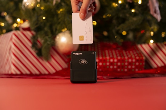 A hand is holding a white credit card above a contactless card reader. The background features a decorated Christmas tree with lights and wrapped gifts in red and white patterns.