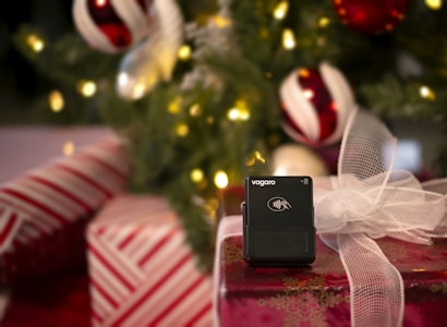 A black wireless payment terminal by Vagaro is placed on a wrapped gift box with a white mesh bow. The background features a decorated Christmas tree with illuminated lights and ornaments in red, white, and green colors.