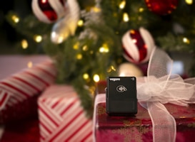 A black wireless payment terminal by Vagaro is placed on a wrapped gift box with a white mesh bow. The background features a decorated Christmas tree with illuminated lights and ornaments in red, white, and green colors.