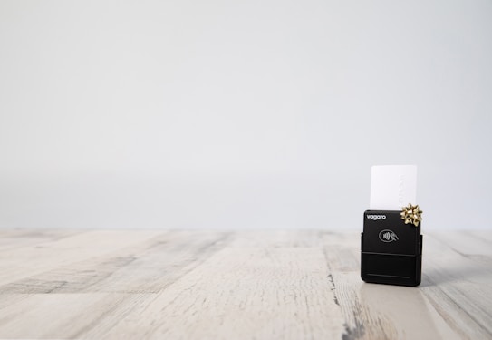 A small black contactless card reader is placed on a light wooden table. The reader has a white card inserted into it and is adorned with a small gold bow. The background is plain and minimalistic, giving a professional and clean appearance.