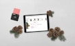 A tablet displaying a Christmas-themed promotion is placed on a light surface alongside pine cones and evergreen branches. A black payment card reader with a pink card inserted is positioned to the left.