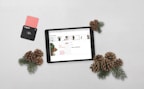 A tablet displaying a Christmas-themed promotion is placed on a light surface alongside pine cones and evergreen branches. A black payment card reader with a pink card inserted is positioned to the left.