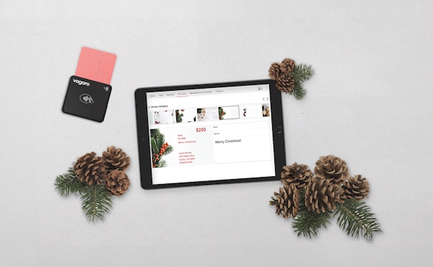 A tablet displaying a Christmas-themed promotion is placed on a light surface alongside pine cones and evergreen branches. A black payment card reader with a pink card inserted is positioned to the left.