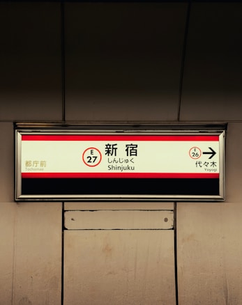 A subway station sign indicates directions in Japanese and English for Shinjuku, with nearby stops named Tochomae and Yoyogi. The sign features a red border and directional arrows, set against a muted background.