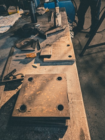 A workshop table cluttered with metal tools and parts, including a large rusted metal plate with holes, various smaller pieces of metal, a screwdriver with an orange handle, and a large file or chisel. The background is dimly lit, suggesting an industrial setting.