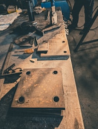 A workshop table cluttered with metal tools and parts, including a large rusted metal plate with holes, various smaller pieces of metal, a screwdriver with an orange handle, and a large file or chisel. The background is dimly lit, suggesting an industrial setting.