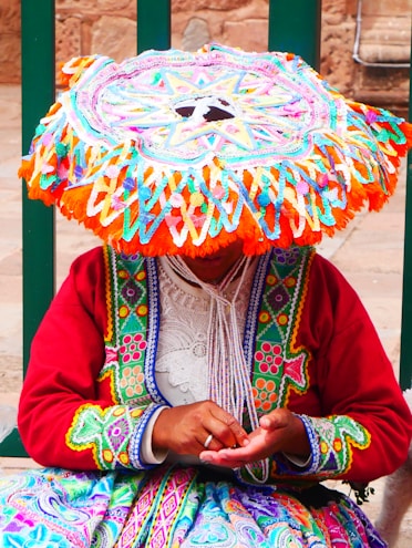 A person wearing vibrant and colorful traditional clothing, including a large, intricately patterned hat that covers their face. The individual's attire features vibrant embroidery in multiple colors like red, green, orange, and blue, and they are holding something in their hands.