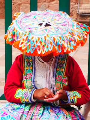 A person wearing vibrant and colorful traditional clothing, including a large, intricately patterned hat that covers their face. The individual's attire features vibrant embroidery in multiple colors like red, green, orange, and blue, and they are holding something in their hands.