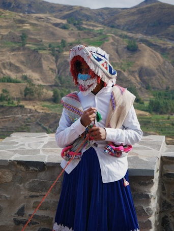 A person dressed in traditional Andean attire stands against a backdrop of rugged mountainous terrain. The colorful costume includes a fringed and beaded mask, a white shirt, and a richly decorated shawl. The person holds a string with one hand while their face is mostly covered by the ornate mask.