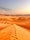 A vast desert landscape with tall sand dunes under a clear sky. There are visible footprints along the ridge of one of the dunes, and vehicles along with tents can be seen in the distance, suggesting a campsite.