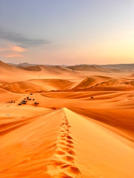A vast desert landscape with tall sand dunes under a clear sky. There are visible footprints along the ridge of one of the dunes, and vehicles along with tents can be seen in the distance, suggesting a campsite.