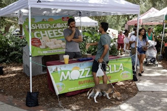 A market stall is set up under a white tent, displaying signs for sheep, goat cheese, and mozzarella. Several people are present; one man in a gray shirt stands behind the table, another in a green shirt with a dog on a leash is talking to him. A woman with long hair in a blue and white outfit walks by. The setting appears to be an outdoor market with trees and other stalls in the background.