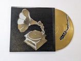 A stylized image of a gramophone painted in shades of gold and white against a dark background. Beside it, a golden vinyl record with a stylized black snake design is partially visible.