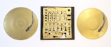 Two gold vinyl records are placed on either side of a square canvas featuring an abstract design resembling a DJ mixer. The records and the central artwork both include detailed turntable tonearms painted in black.