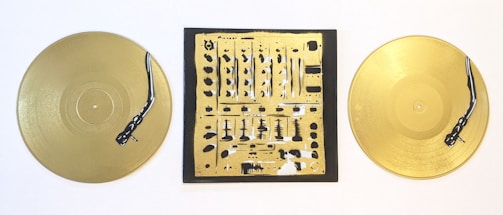 Two gold vinyl records are placed on either side of a square canvas featuring an abstract design resembling a DJ mixer. The records and the central artwork both include detailed turntable tonearms painted in black.