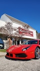 A bright red sports car is parked in front of a dealership building. The building has light-colored siding with a red and white sign. There are some trees with sparse leaves nearby, and the sky is clear and blue.