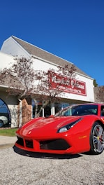 A bright red sports car is parked in front of a dealership building. The building has light-colored siding with a red and white sign. There are some trees with sparse leaves nearby, and the sky is clear and blue.