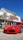 A bright red sports car is parked in front of a dealership building. The building has light-colored siding with a red and white sign. There are some trees with sparse leaves nearby, and the sky is clear and blue.