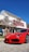 A bright red sports car is parked in front of a dealership building. The building has light-colored siding with a red and white sign. There are some trees with sparse leaves nearby, and the sky is clear and blue.