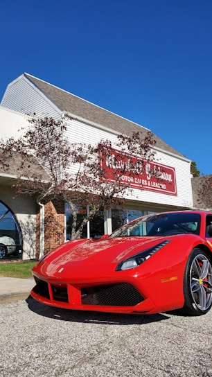 A bright red sports car is parked in front of a dealership building. The building has light-colored siding with a red and white sign. There are some trees with sparse leaves nearby, and the sky is clear and blue.