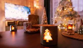 A cozy room decorated for the holidays, featuring a lit candle with a Christmas tree cut-out as the centerpiece on a table. In the background, there is a warmly lit Christmas tree adorned with ornaments, and a television displaying a bright scene. The atmosphere is festive and inviting, with other decorative elements like small Christmas trees and gift-wrapped presents.