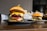 A juicy cheeseburger with a glossy bun, layered with melted cheese, a beef patty, fresh lettuce, tomato, pickles, and red onion. A skewer holds the burger together. In the background, there is another burger on a plate, slightly out of focus. A side of fries wrapped in paper is also visible.