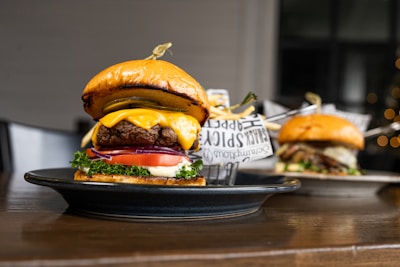 A juicy cheeseburger with a glossy bun, layered with melted cheese, a beef patty, fresh lettuce, tomato, pickles, and red onion. A skewer holds the burger together. In the background, there is another burger on a plate, slightly out of focus. A side of fries wrapped in paper is also visible.