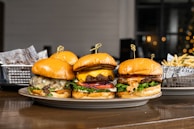 Three gourmet burgers are displayed on a plate, each with different toppings including cheese, lettuce, tomatoes, and sauces. Behind the burgers, there is a side of french fries in a metal basket lined with decorative paper. The setting appears to be a casual dining environment with a blurred background that features some warm lighting.