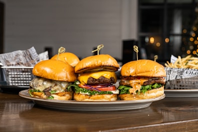 Three gourmet burgers are displayed on a plate, each with different toppings including cheese, lettuce, tomatoes, and sauces. Behind the burgers, there is a side of french fries in a metal basket lined with decorative paper. The setting appears to be a casual dining environment with a blurred background that features some warm lighting.