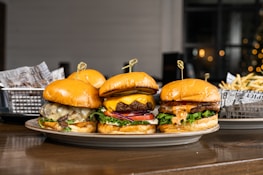 Three gourmet burgers are displayed on a plate, each with different toppings including cheese, lettuce, tomatoes, and sauces. Behind the burgers, there is a side of french fries in a metal basket lined with decorative paper. The setting appears to be a casual dining environment with a blurred background that features some warm lighting.