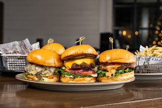 Three gourmet burgers are displayed on a plate, each with different toppings including cheese, lettuce, tomatoes, and sauces. Behind the burgers, there is a side of french fries in a metal basket lined with decorative paper. The setting appears to be a casual dining environment with a blurred background that features some warm lighting.