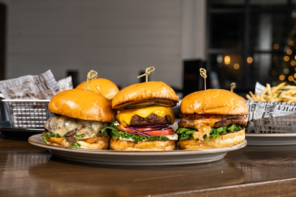 Three gourmet burgers are displayed on a plate, each with different toppings including cheese, lettuce, tomatoes, and sauces. Behind the burgers, there is a side of french fries in a metal basket lined with decorative paper. The setting appears to be a casual dining environment with a blurred background that features some warm lighting.
