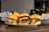 Three gourmet burgers are displayed on a plate, each with different toppings including cheese, lettuce, tomatoes, and sauces. Behind the burgers, there is a side of french fries in a metal basket lined with decorative paper. The setting appears to be a casual dining environment with a blurred background that features some warm lighting.