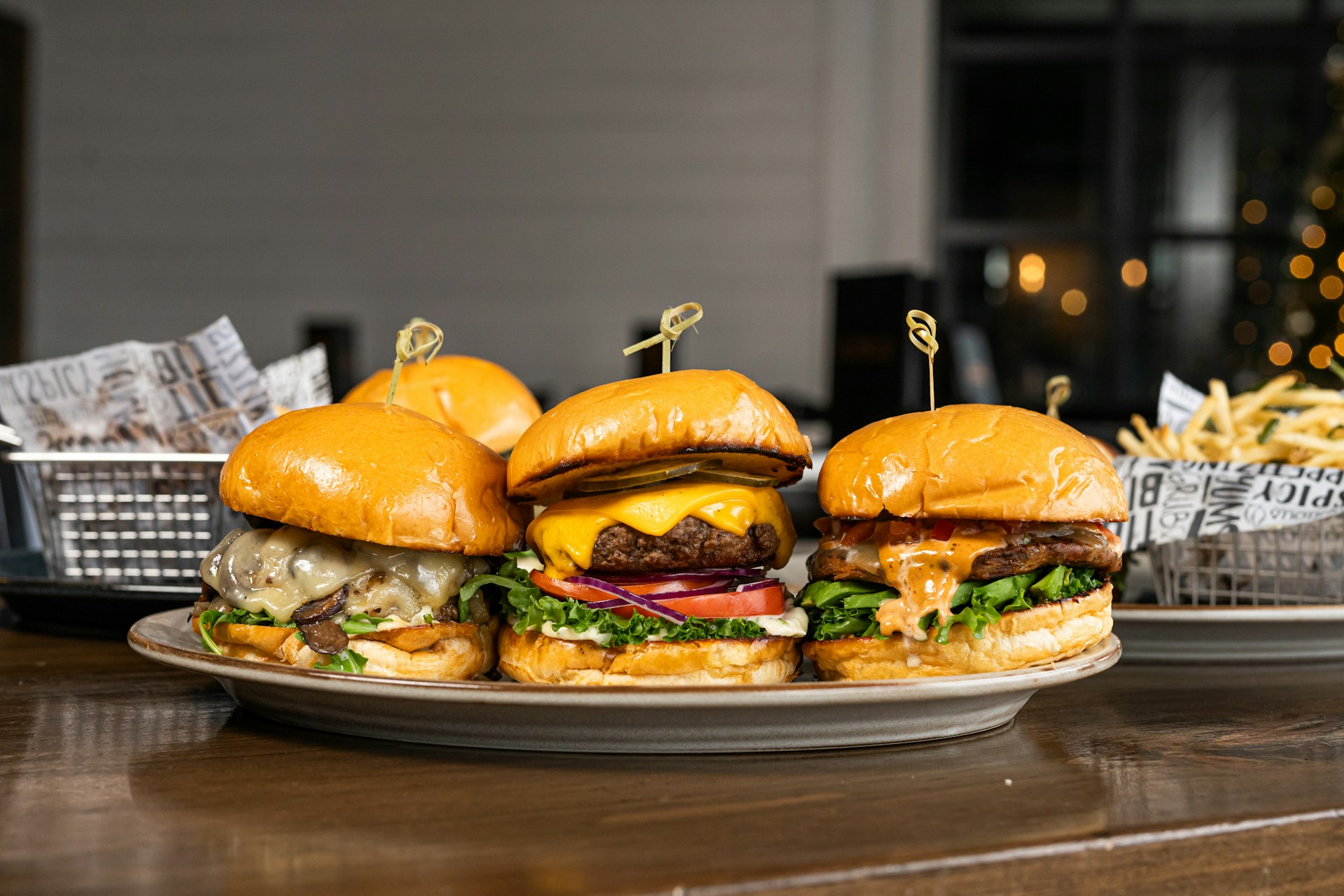 Three gourmet burgers are displayed on a plate, each with different toppings including cheese, lettuce, tomatoes, and sauces. Behind the burgers, there is a side of french fries in a metal basket lined with decorative paper. The setting appears to be a casual dining environment with a blurred background that features some warm lighting.