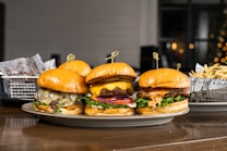 Three gourmet burgers are displayed on a plate, each with different toppings including cheese, lettuce, tomatoes, and sauces. Behind the burgers, there is a side of french fries in a metal basket lined with decorative paper. The setting appears to be a casual dining environment with a blurred background that features some warm lighting.