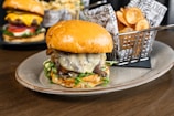 A gourmet burger with a toasted bun, melted cheese, caramelized onions, and greens is served on an oval plate. In the background, there is a basket filled with crispy potato chips and another burger with fresh toppings like lettuce, tomato, and cheese.