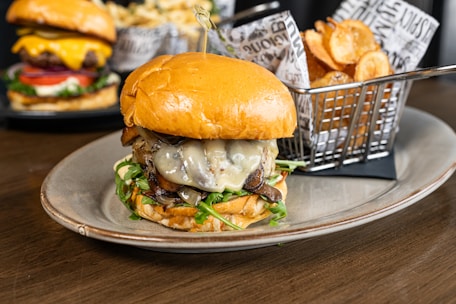 A gourmet burger with a toasted bun, melted cheese, caramelized onions, and greens is served on an oval plate. In the background, there is a basket filled with crispy potato chips and another burger with fresh toppings like lettuce, tomato, and cheese.