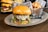 A gourmet burger with a toasted bun, melted cheese, caramelized onions, and greens is served on an oval plate. In the background, there is a basket filled with crispy potato chips and another burger with fresh toppings like lettuce, tomato, and cheese.
