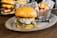 A gourmet burger with a toasted bun, melted cheese, caramelized onions, and greens is served on an oval plate. In the background, there is a basket filled with crispy potato chips and another burger with fresh toppings like lettuce, tomato, and cheese.