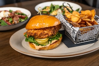 A fried chicken sandwich topped with lettuce and pickles is served on a round plate, accompanied by a metal basket filled with crispy potato chips. In the background, two bowls of salad with a variety of fresh ingredients are placed on a wooden table.