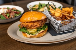 A fried chicken sandwich topped with lettuce and pickles is served on a round plate, accompanied by a metal basket filled with crispy potato chips. In the background, two bowls of salad with a variety of fresh ingredients are placed on a wooden table.
