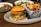 A fried chicken sandwich topped with lettuce and pickles is served on a round plate, accompanied by a metal basket filled with crispy potato chips. In the background, two bowls of salad with a variety of fresh ingredients are placed on a wooden table.