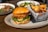 A fried chicken sandwich topped with lettuce and pickles is served on a round plate, accompanied by a metal basket filled with crispy potato chips. In the background, two bowls of salad with a variety of fresh ingredients are placed on a wooden table.