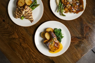 Three plates of food arranged on a wooden table, each featuring grilled chicken, sliced potatoes, and green vegetables. One dish has a rich sauce, while others include grilled asparagus.