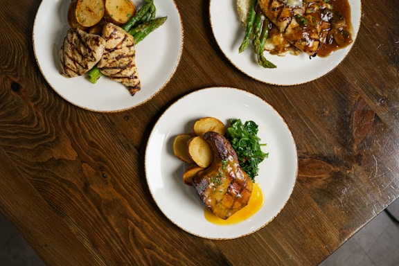 Three plates of food arranged on a wooden table, each featuring grilled chicken, sliced potatoes, and green vegetables. One dish has a rich sauce, while others include grilled asparagus.
