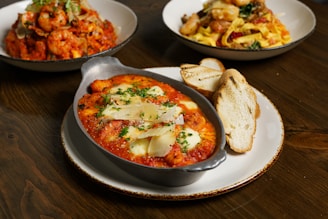 A wooden table displays a main dish of baked gnocchi with rich tomato sauce, melted cheese, and fresh herbs in a rustic metal dish, accompanied by slices of toasted bread on a white plate. In the background, two dishes filled with pasta and shrimp sit, featuring vibrant colors and garnished with greens.